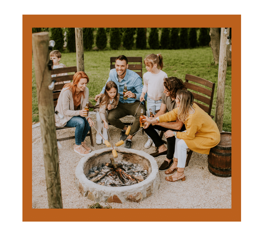 Smiling family roasting marshmallows together around a backyard fire pit on a clear day.
